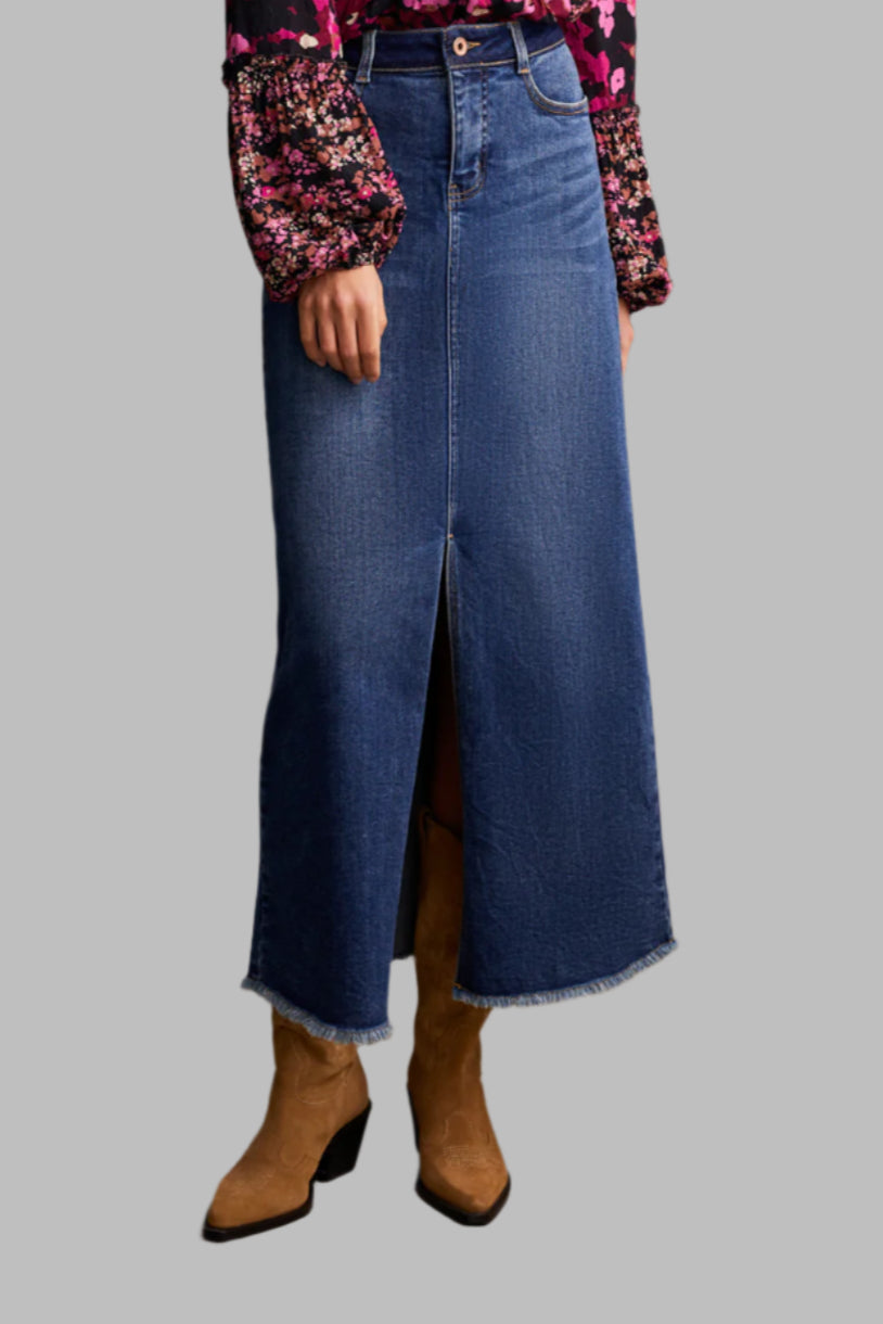A woman standing in a studio setting, wearing a long maxi skirt with 5 pockets, paired with a floral shirt and brown boots.