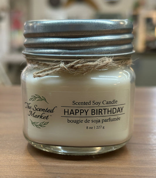 Scented soy candle labeled 'Happy Birthday' on a wooden surface with a blurred indoor background.