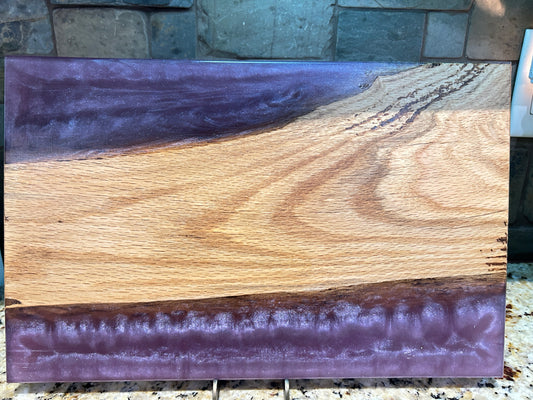 A rectangular wooden board or plaque with a natural live-edge wood grain pattern, featuring a prominent purple resin border on the top and bottom, displayed against a stone tile background.