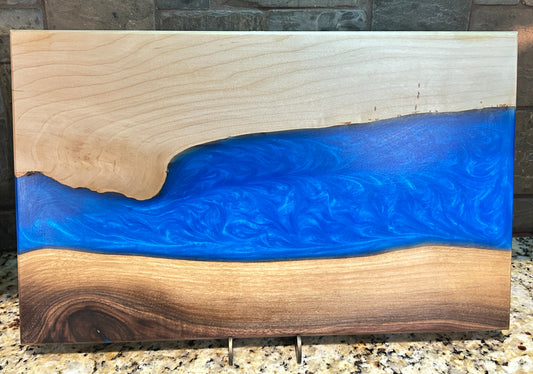 A rectangular wooden board with a vibrant blue resin river design flowing through natural wood grain sections, displayed on a granite countertop against a stone tile backsplash.