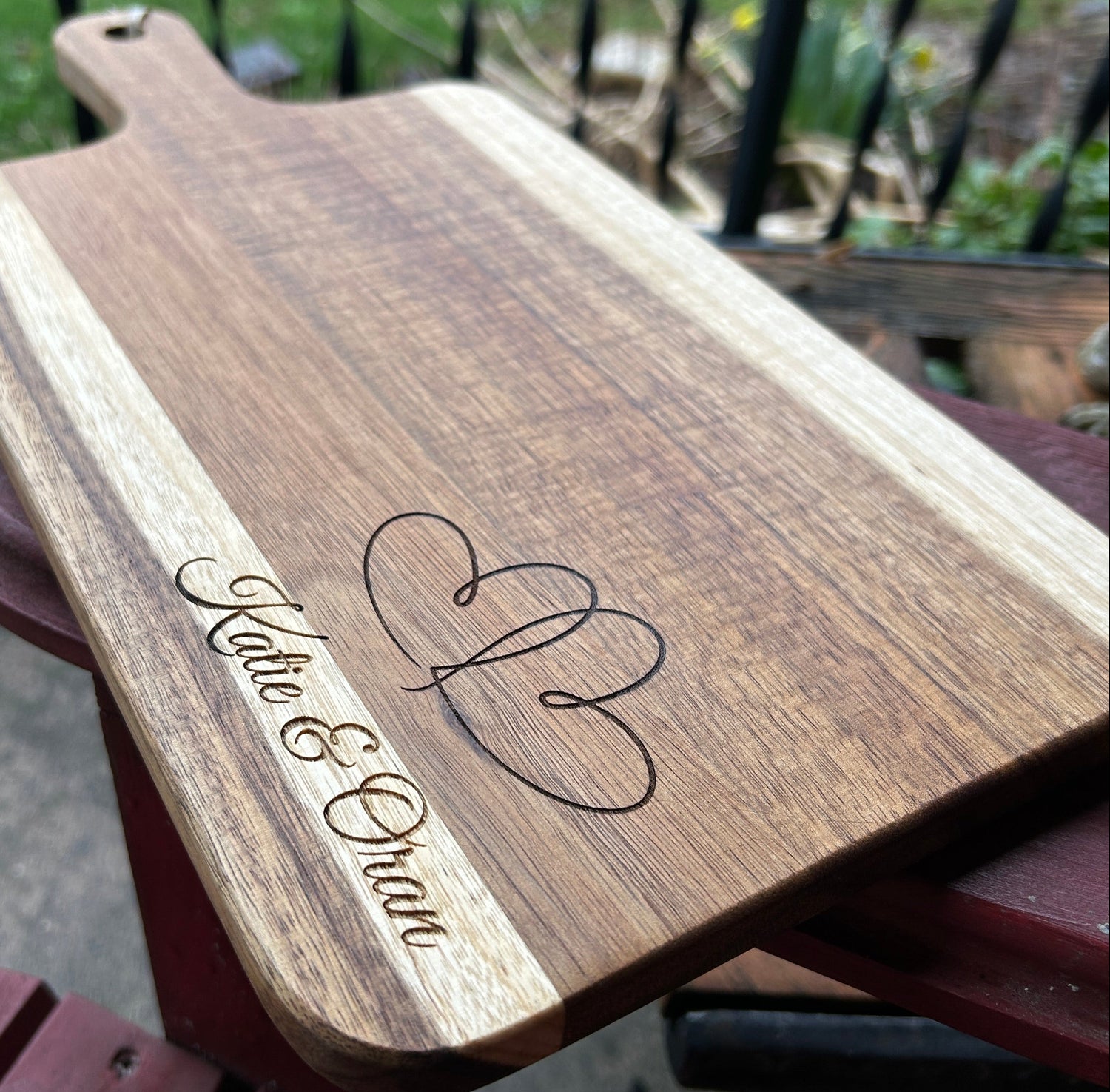 Wooden cutting board with engraved heart design and text, held against a blurred outdoor background.