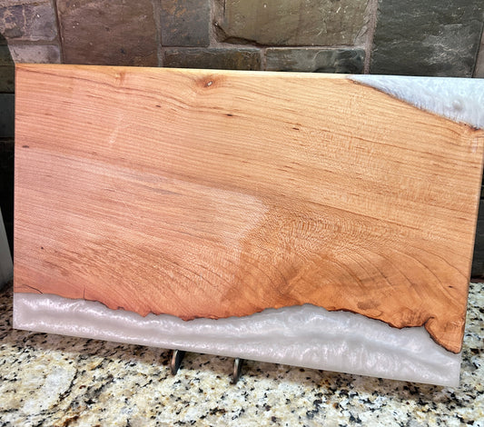 A rectangular live-edge wooden board with a natural grain pattern and a white resin or stone bottom edge, displayed on a granite countertop in front of a stone tile wall.