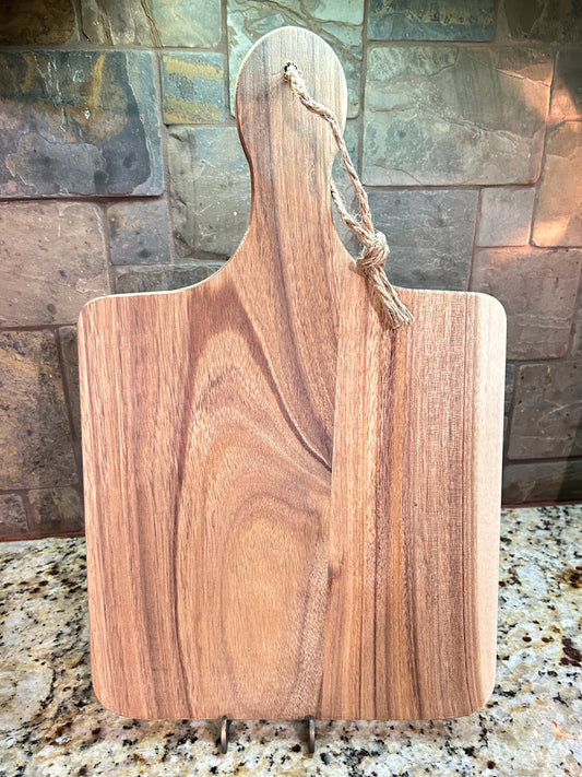 A rectangular wooden cutting board with rounded corners and a handle, shown standing upright on a small metal stand on a granite countertop with a stone tile backsplash behind it.