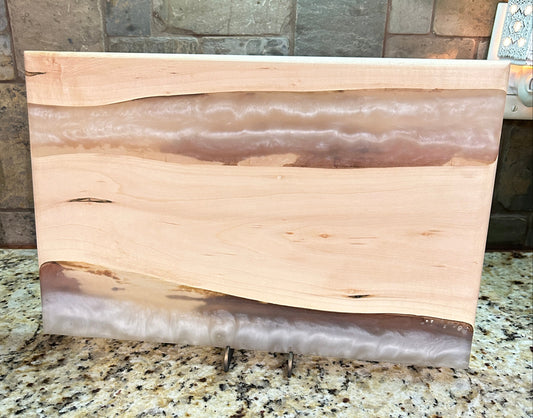A rectangular wooden board with wavy bands of natural wood and agate-like stone inlay, displayed on a granite countertop against a stone tile backsplash.