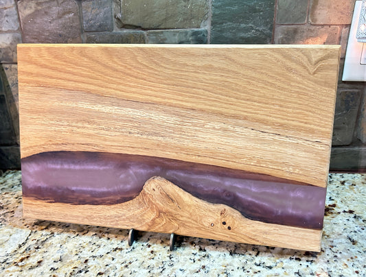 A rectangular wooden board with a natural live edge and a glossy dark purple resin inlay, displayed on a granite countertop in front of a stone tile backsplash.