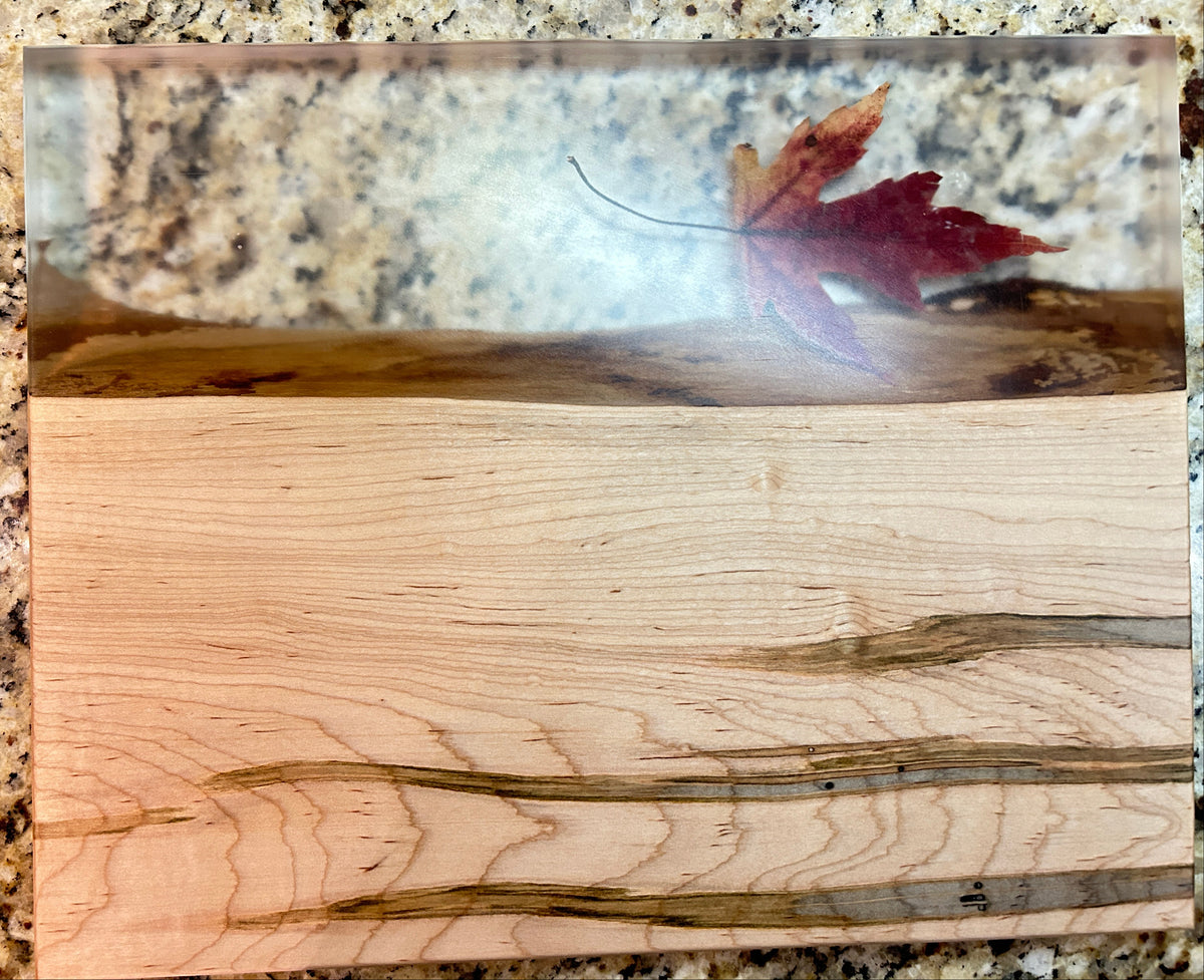 A rectangular wooden board or tray with a split design: one side shows light natural wood grain, and the other side has a glossy clear resin section containing a red autumn leaf. The board is photographed on a speckled granite countertop.