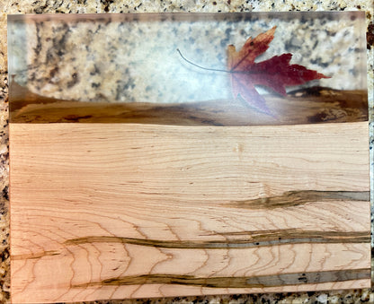 A rectangular wooden board or tray with a split design: one side shows light natural wood grain, and the other side has a glossy clear resin section containing a red autumn leaf. The board is photographed on a speckled granite countertop.