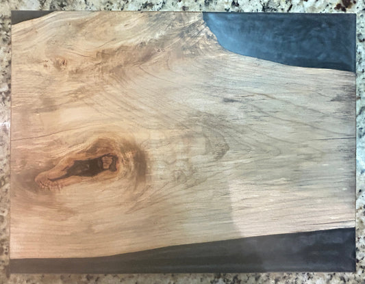 A rectangular wooden board or plaque with a live edge showing natural wood grain, knots, and dark resin accents on the sides, photographed on a speckled granite countertop.