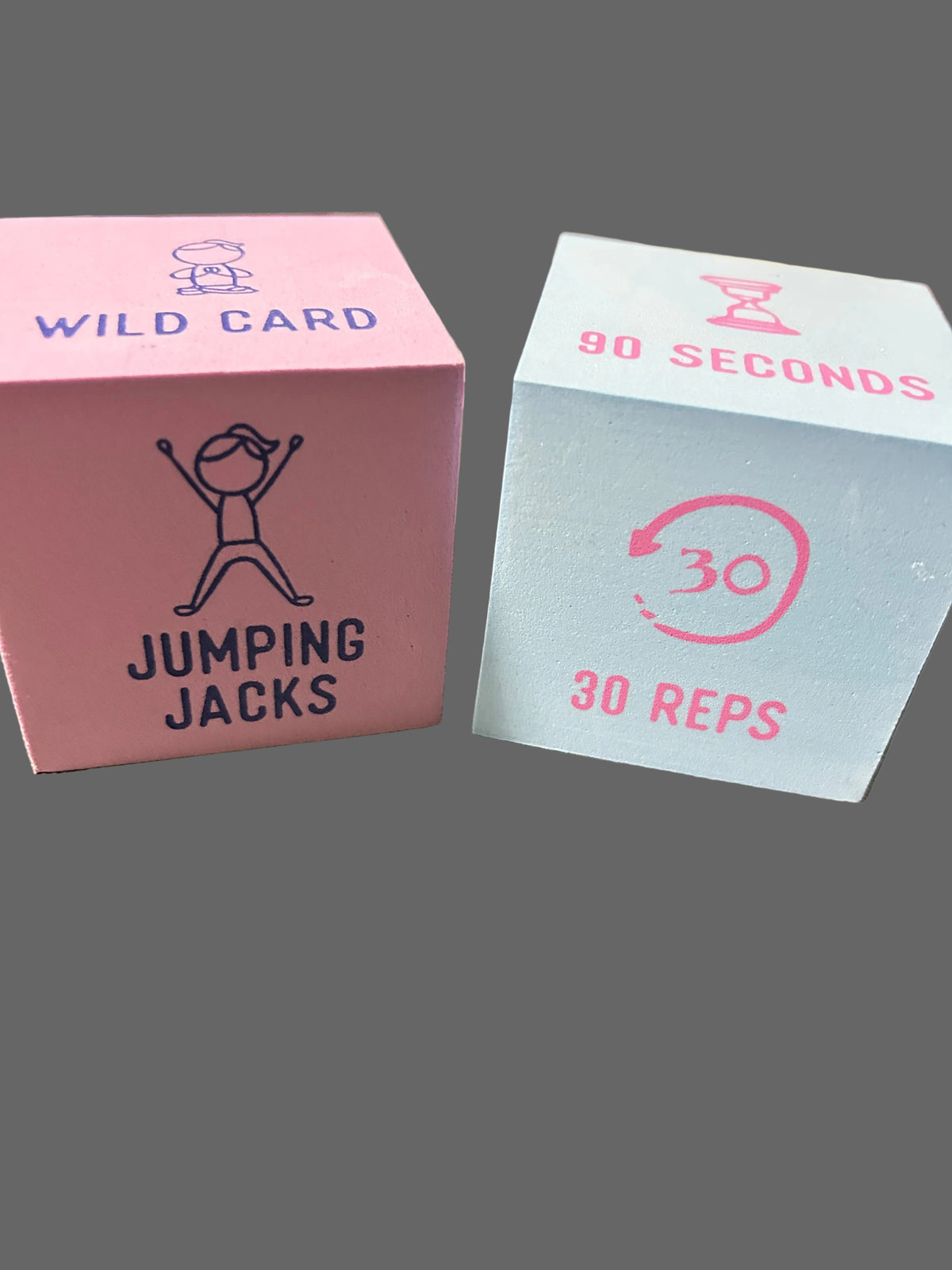 Two pink and light blue fitness tracking cubes on a textured gray fabric surface. The pink cube is labeled 'WILD CARD' and 'JUMPING JACKS', while the light blue cube is labeled '90 SECONDS' and '30 REPS'.